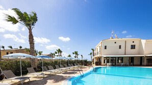 A pool area with lounge chairs and umbrellas, a palm tree, and a building with a tiled roof. - Resort Santa Maria (Marsala)