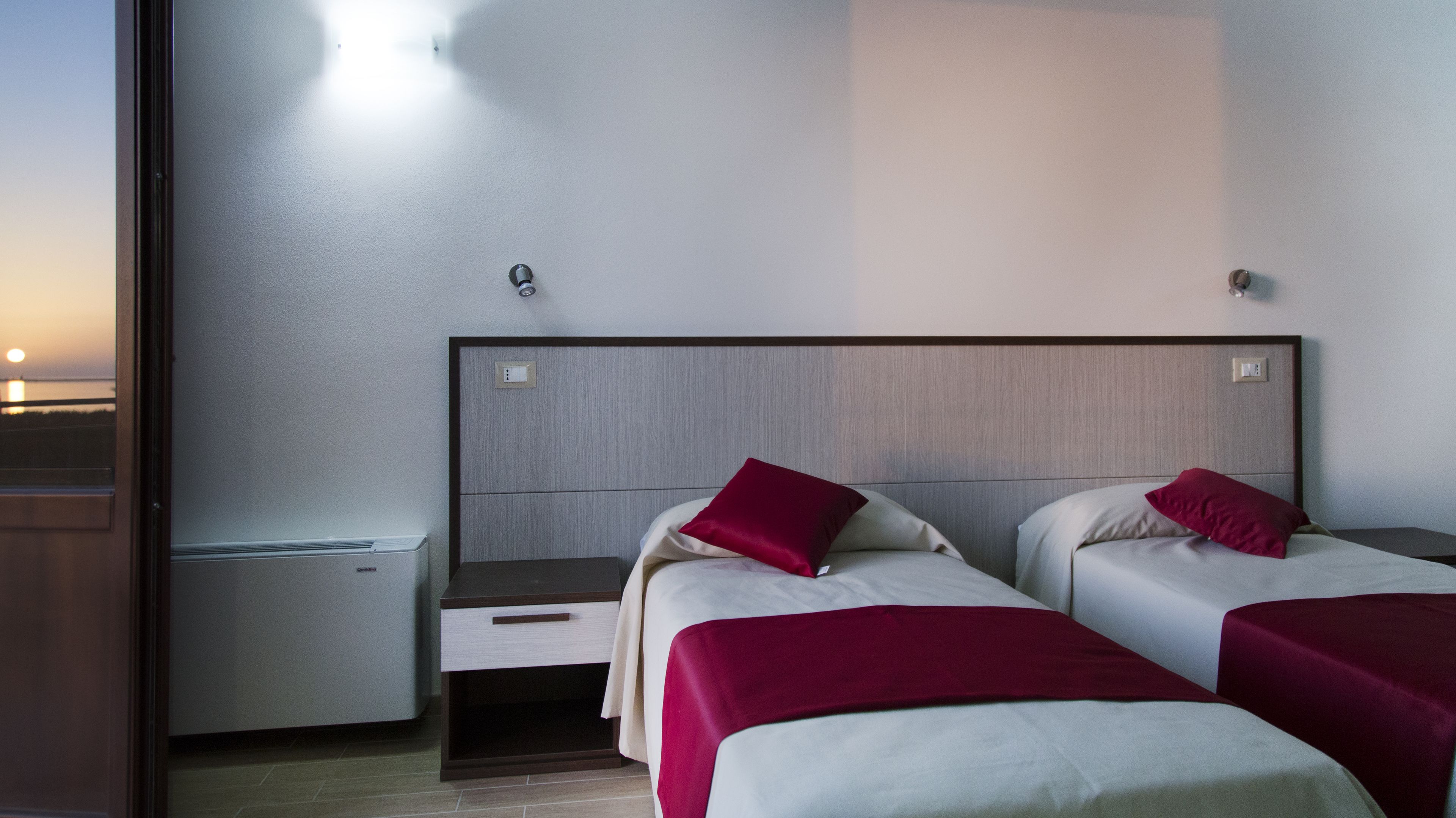 A hotel room with two beds, a nightstand, a small table, and a window with a view of the sunset.