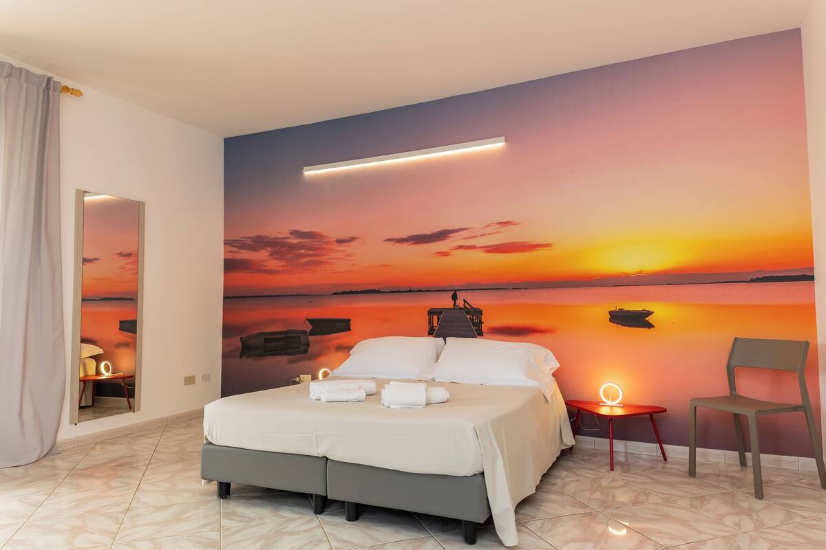 A bedroom with a large bed, a nightstand, a chair, and a wall mural of a sunset over water.
