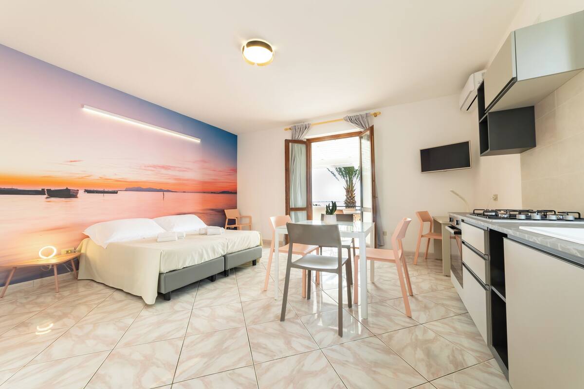 A modern bedroom with a bed, bedside table, and a view of a sunset through a window.