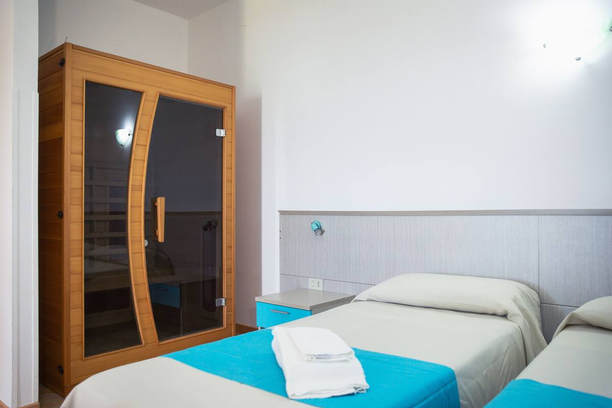 A hotel room with a large wardrobe, two beds with blue bedding, and a small bedside table.