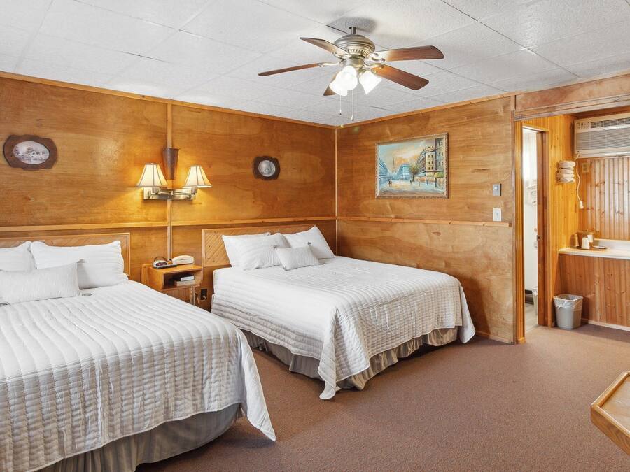 Hotelkamer met twee bedden, een plafondventilator, een schilderij, een nachtkastje, een lamp en een badkamer met een wastafel en spiegel.