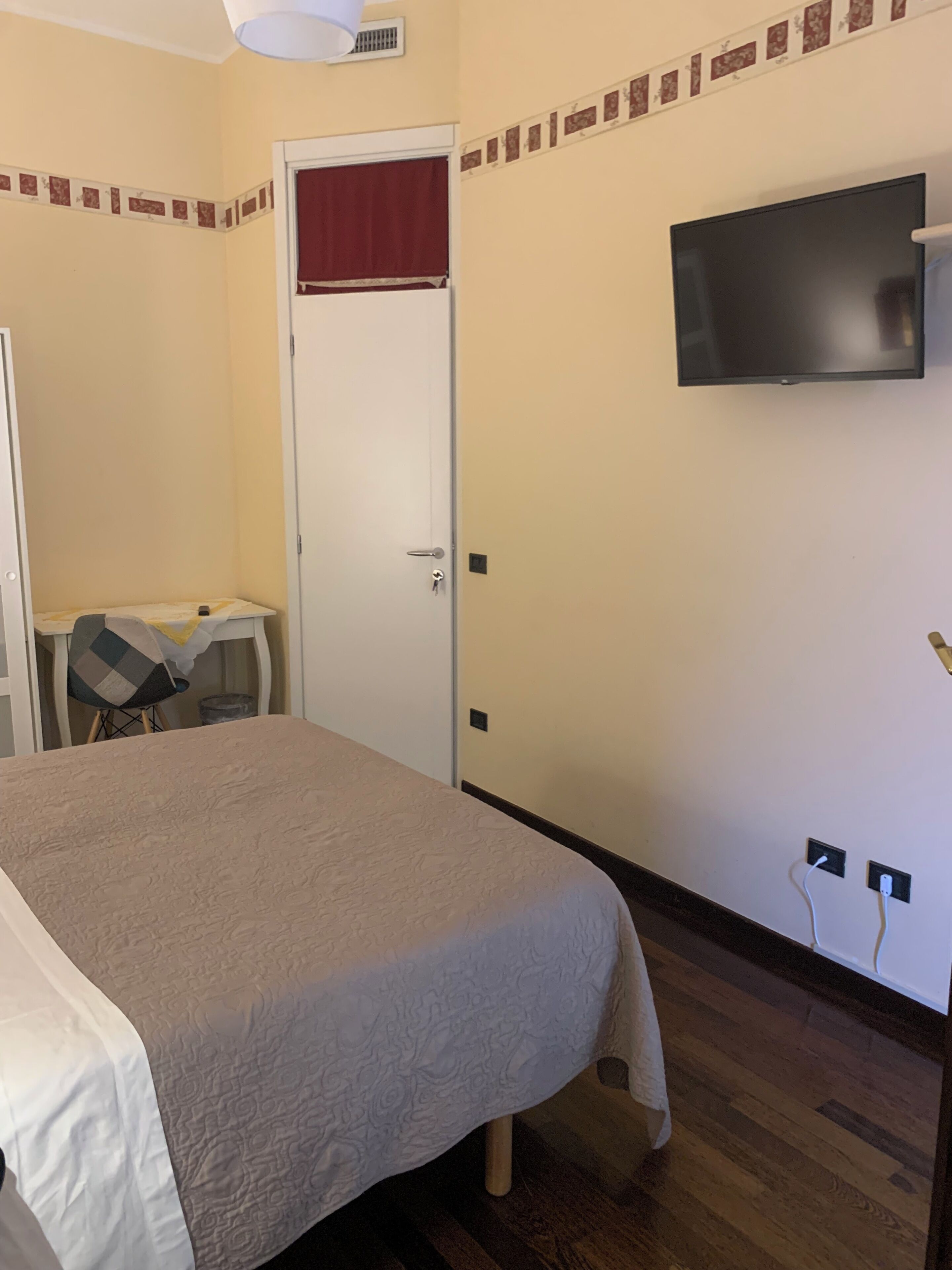 Ett sovrum med en säng, en tv som är monterad på väggen, ett skrivbord med en stol och en dörr med en röd gardin.