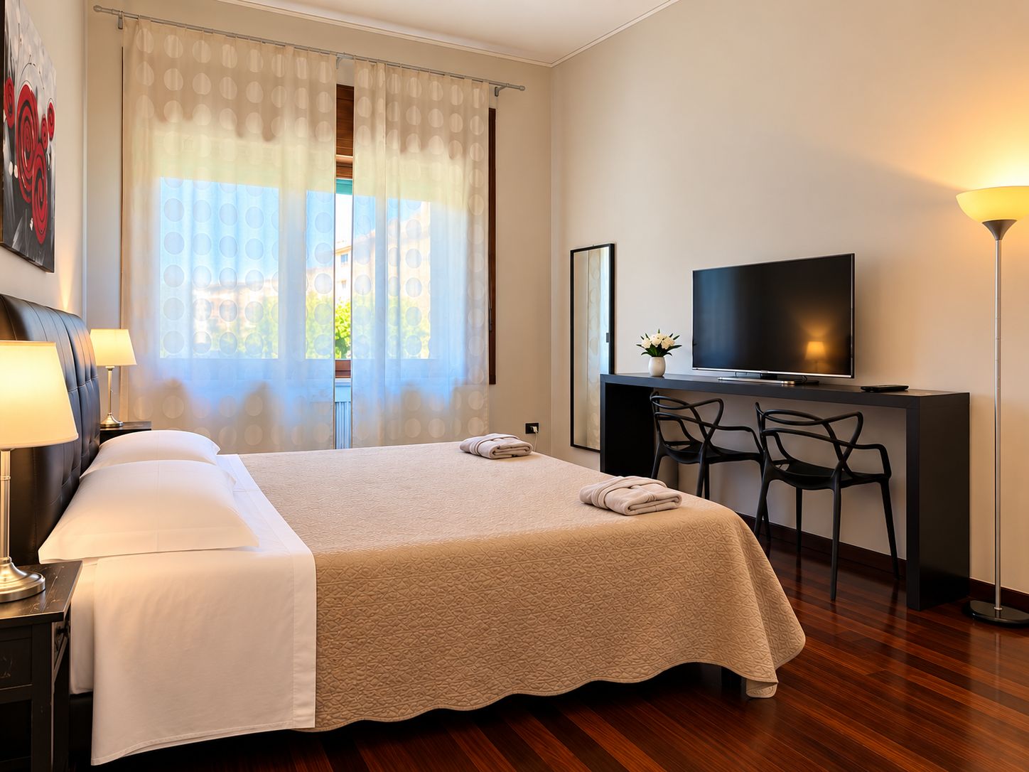 Ein Hotelzimmer mit einem Bett, einem Schreibtisch mit Fernseher, zwei Stühlen, einer Schreibtischlampe und einem Fenster mit Vorhängen.