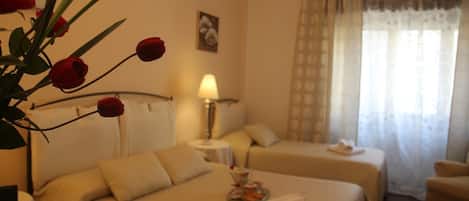 A hotel room with two beds, a bedside table with a lamp, a small table with a tray of food, and a large window with curtains.