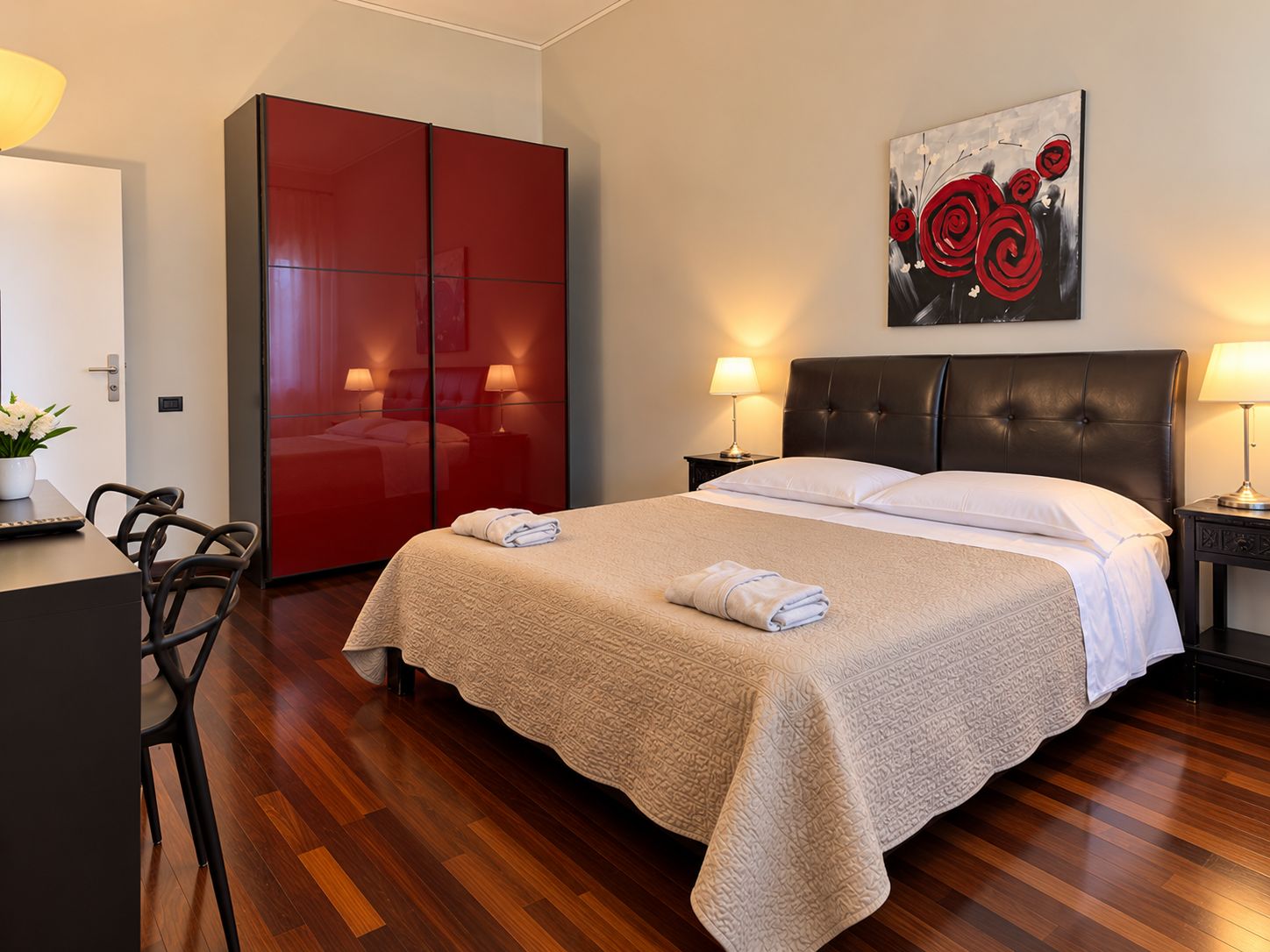 A hotel room with a bed, a red wardrobe, a painting, and a desk with a chair.