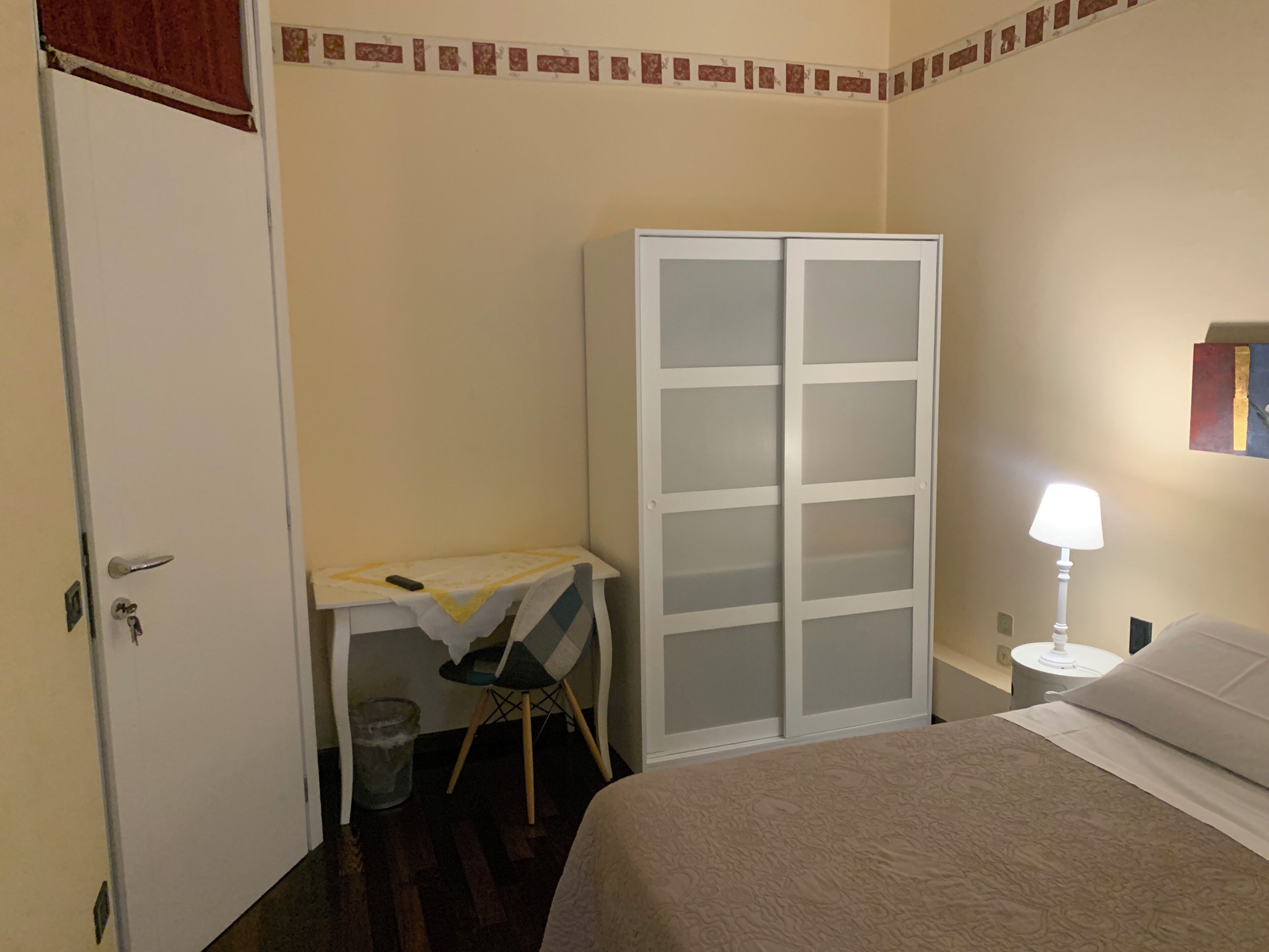 A hotel room with a bed, a desk with a chair, a wardrobe, a trash bin, and a nightstand with a lamp.