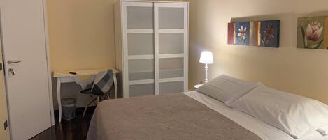 A bedroom with a bed, a desk, a wardrobe, and a trash bin.