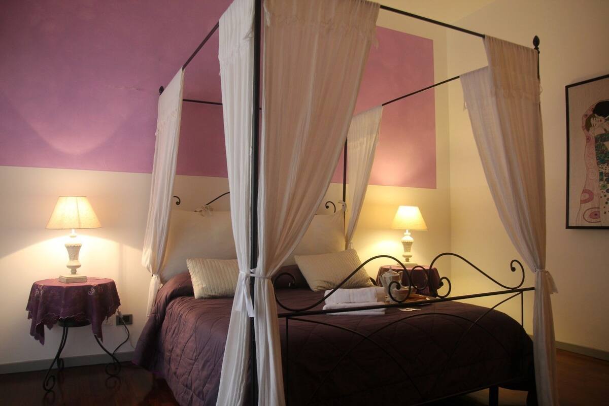 Een bed met hemelbed, twee nachtkastjes met lampen en een ingelijst schilderij aan de muur.
