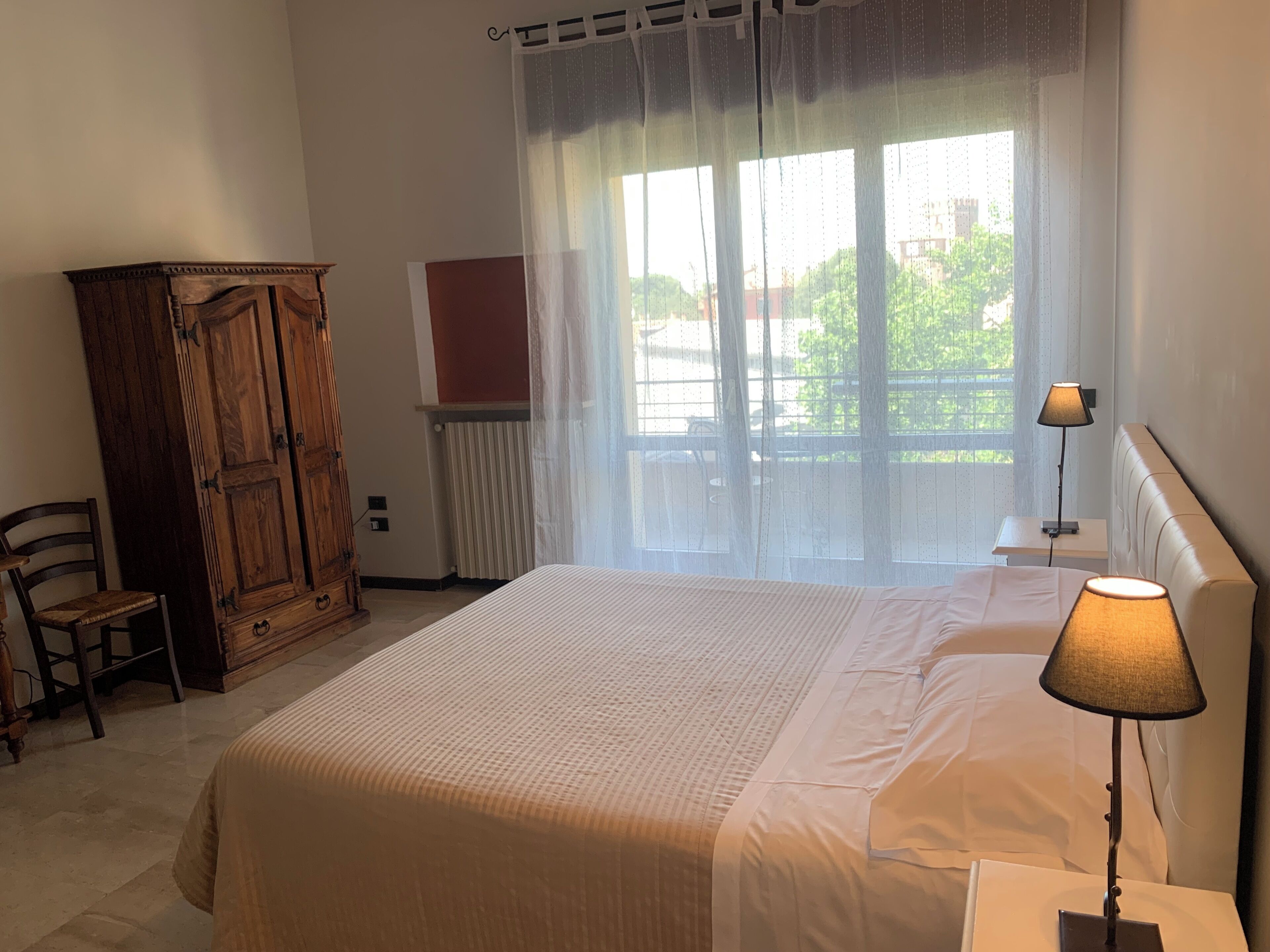 Un dormitorio con cama, armario de madera, silla, radiador, ventana con cortinas y lámpara de noche.