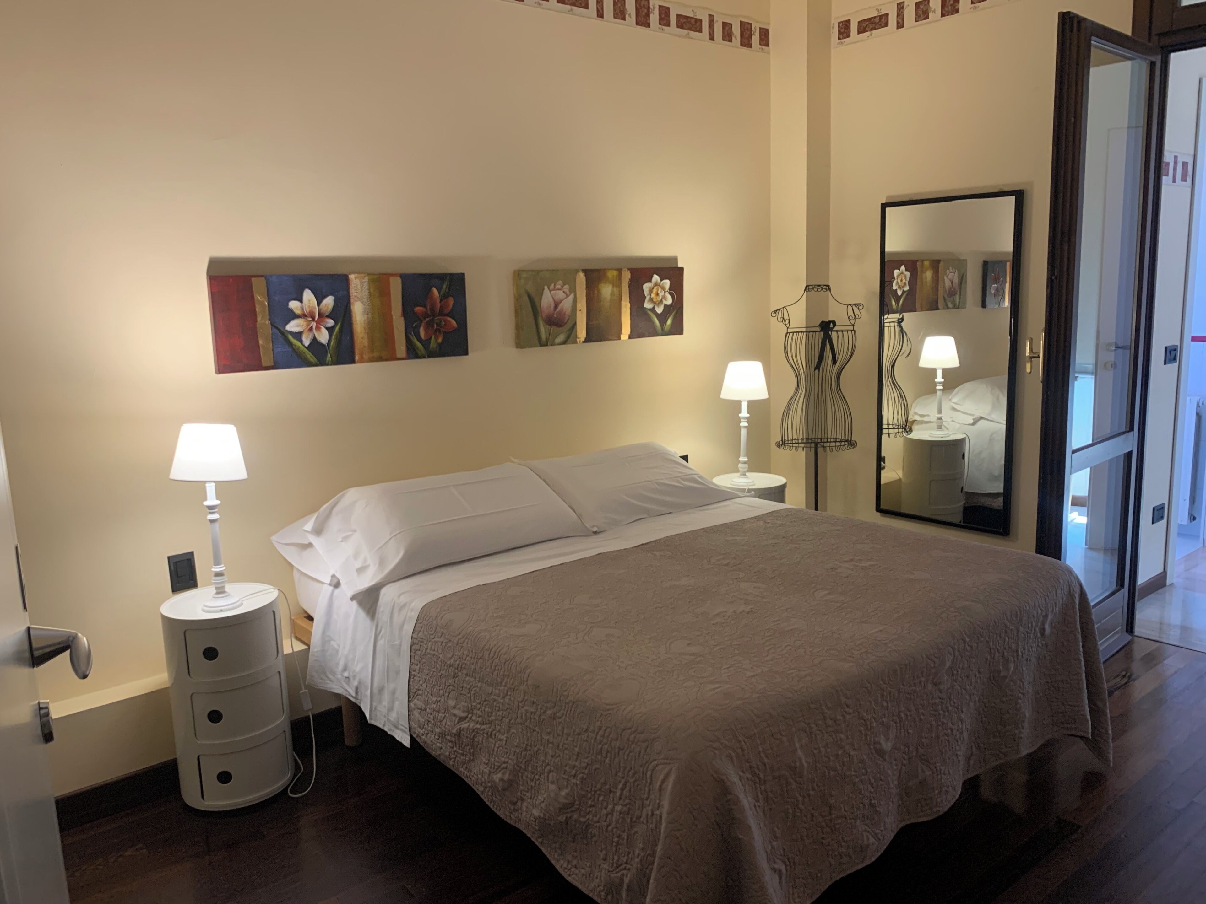 A bedroom with a bed, two bedside tables with lamps, a mirror, and floral artwork on the wall.