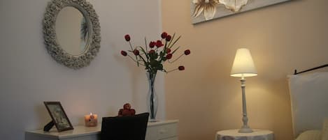 Un dormitorio con cama, mesita de noche con lámpara, cómoda, espejo con marco floral, un jarrón con flores rojas y un cuadro en la pared.