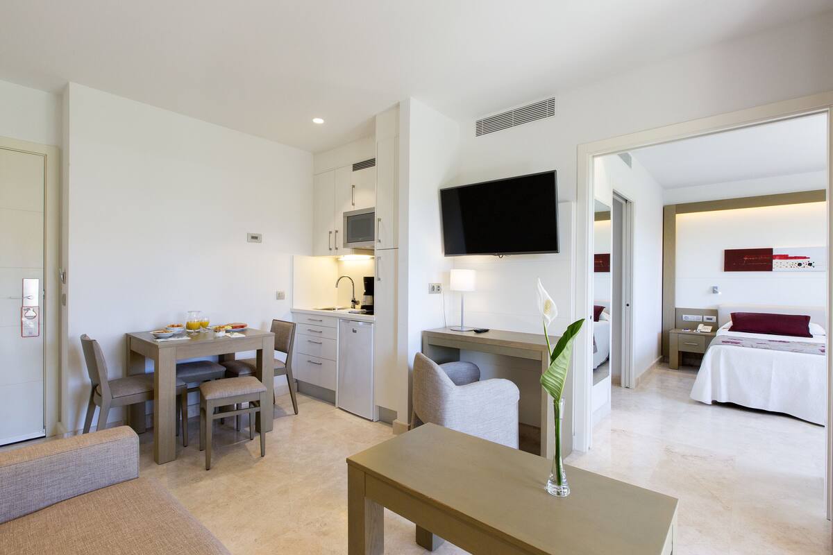 A hotel room with a dining area, kitchenette, TV, bed, and a bathroom visible through an open door.