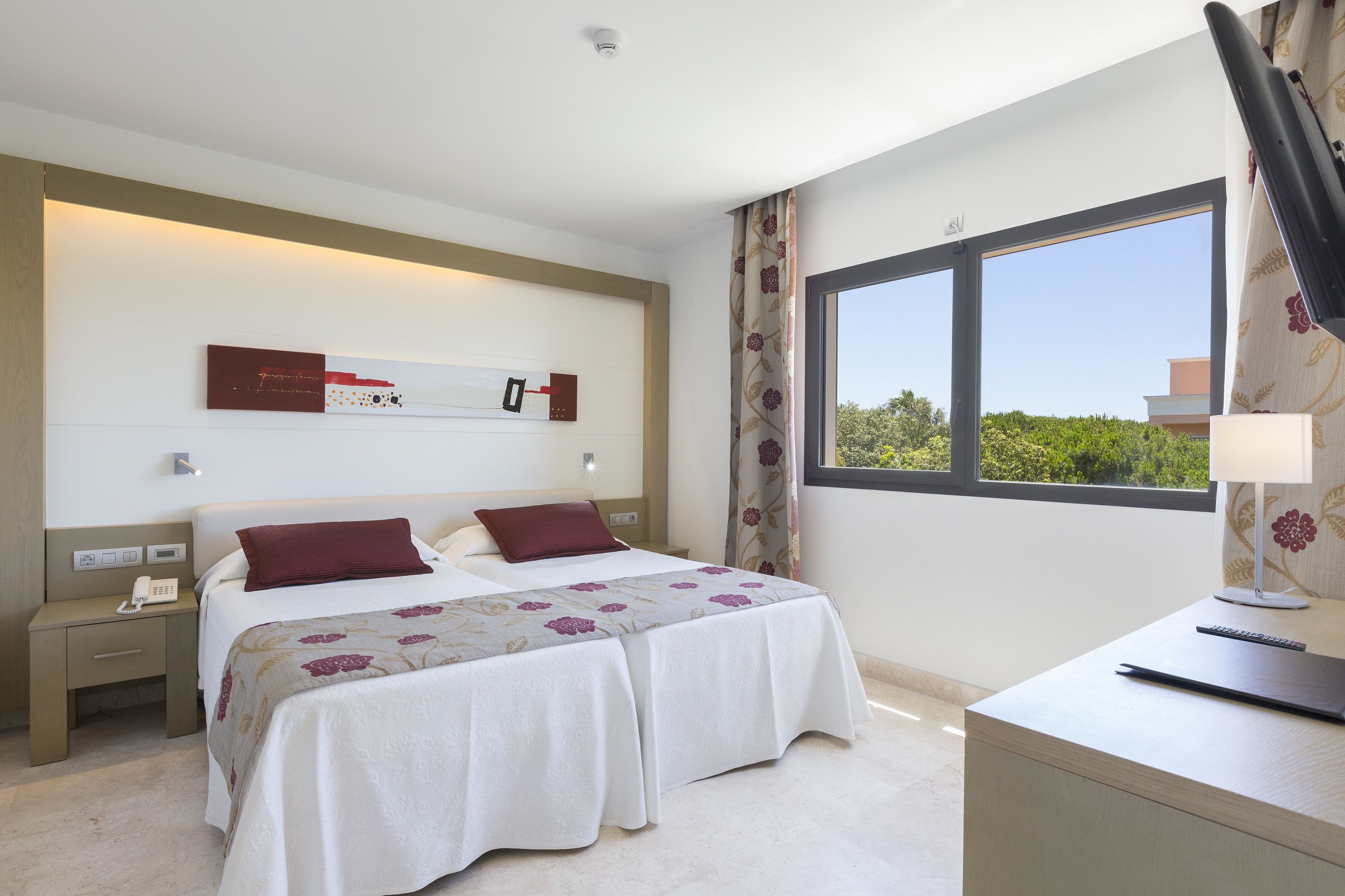 Habitación de hotel con dos camas, un escritorio, una lámpara y una ventana con vistas a zonas verdes.