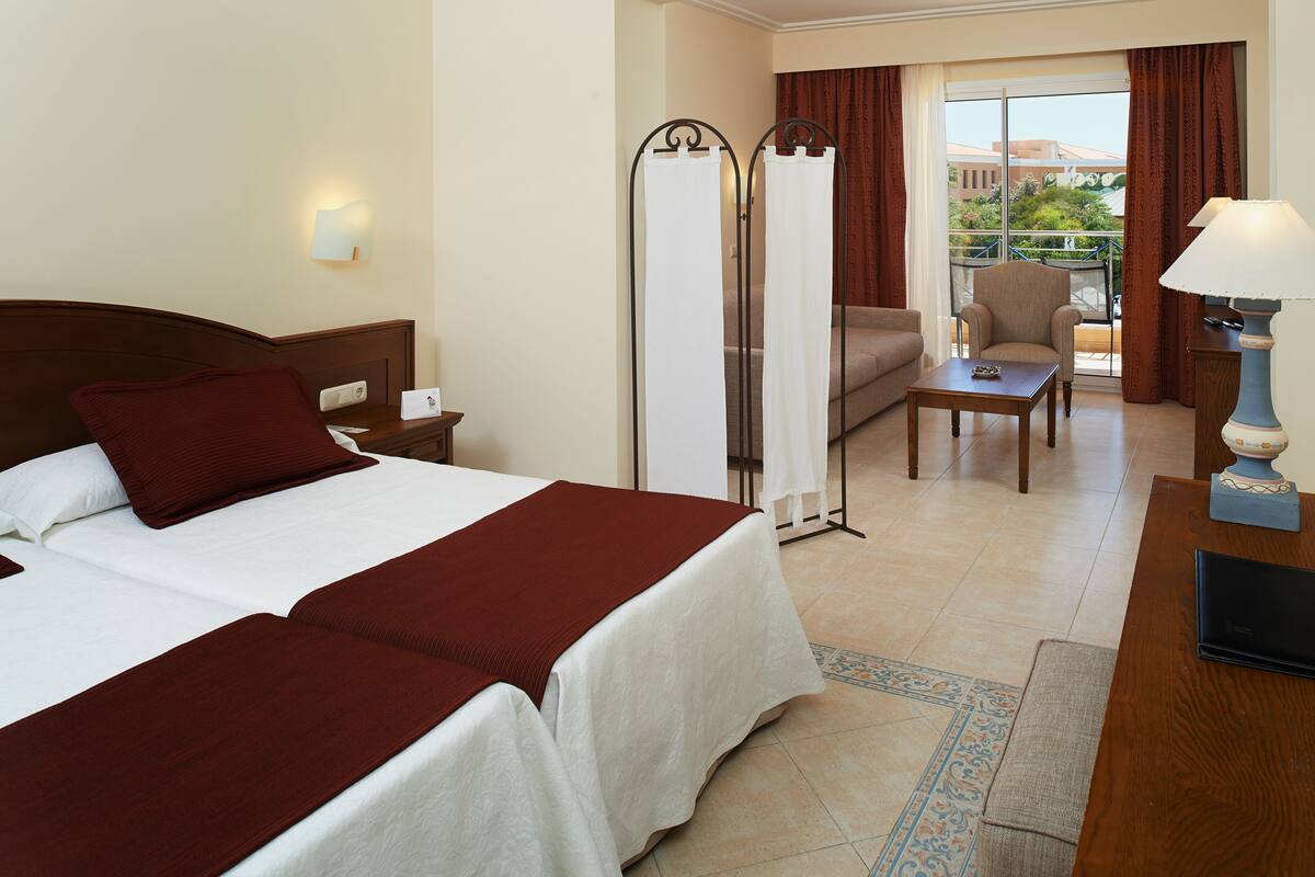 A hotel room with a large bed, a sofa, a small table, a lamp, and a view of the outdoors through a sliding door.