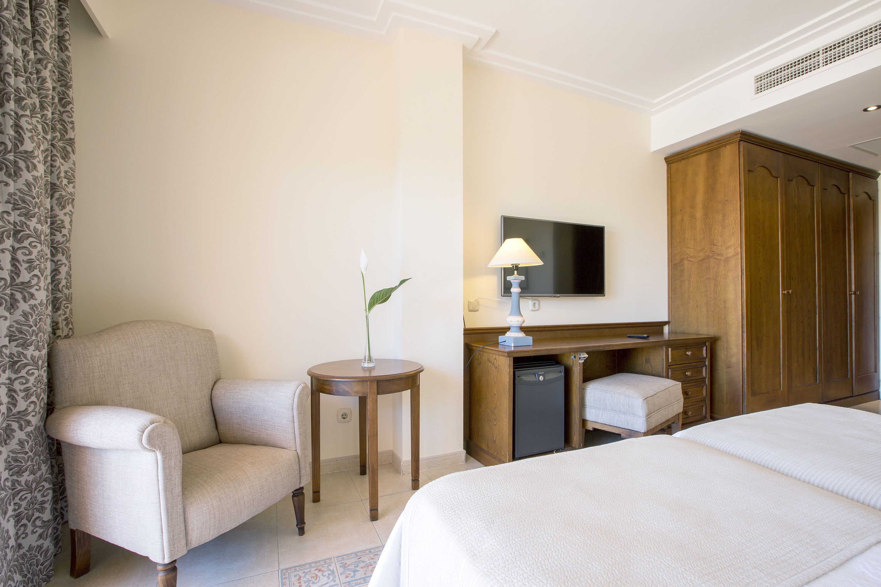 A hotel room with a bed, a chair, a small table with a lamp, a television, a wardrobe, and a desk with a refrigerator.