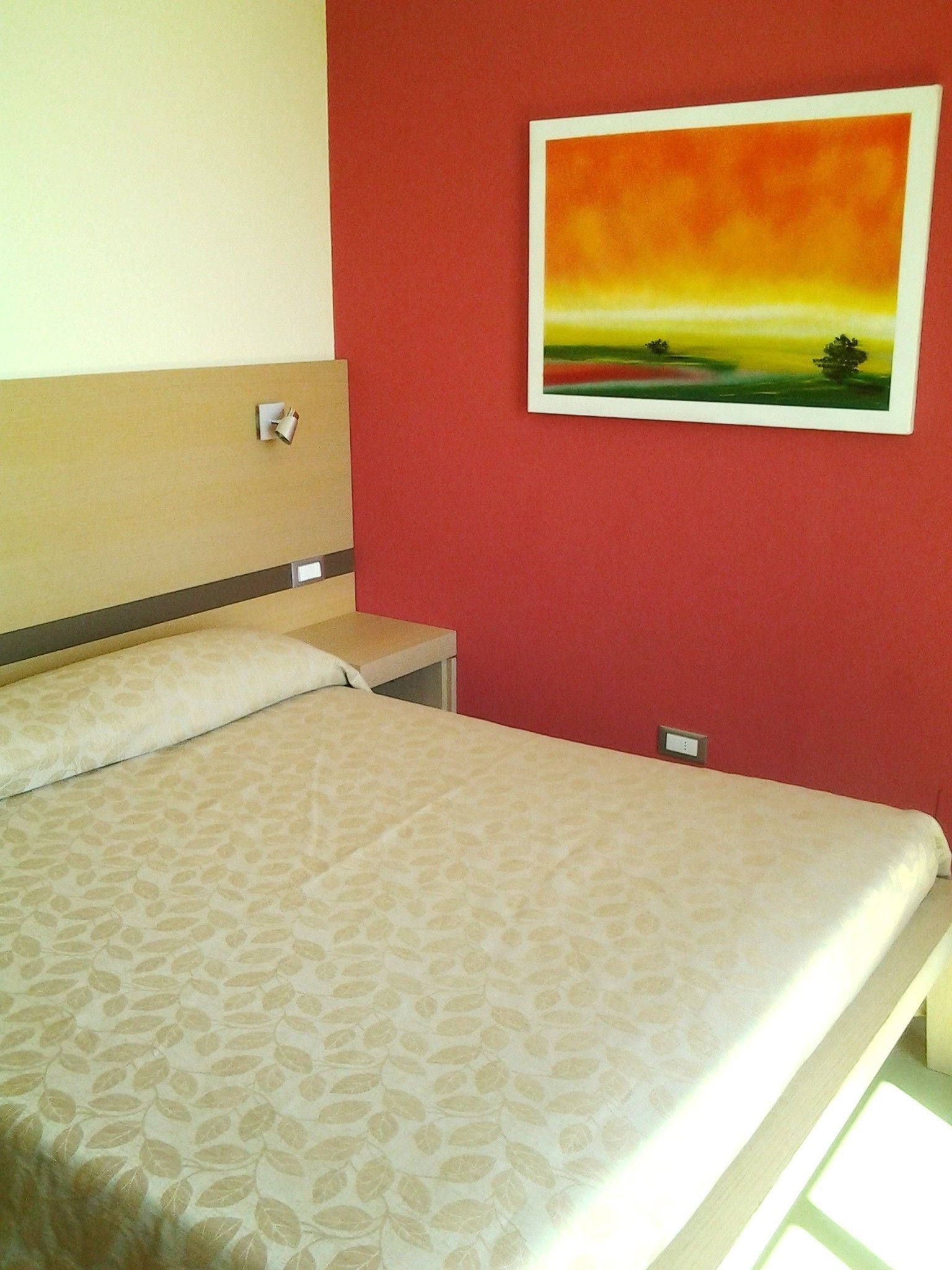 Una habitación de hotel con cama, mesita de noche, lámpara de pared y un cuadro con un paisaje de atardecer.