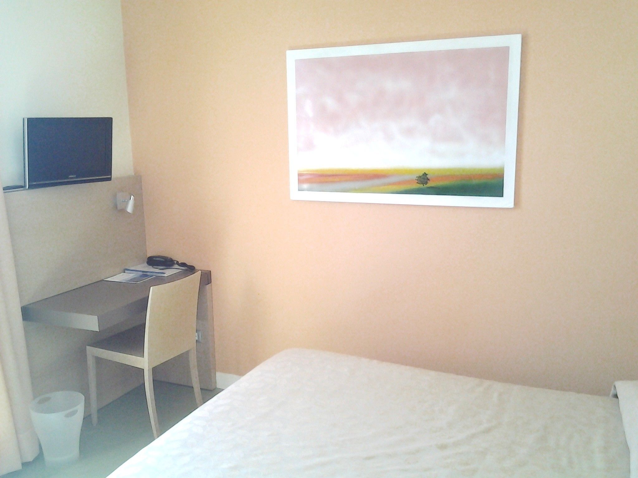 A hotel room with a bed, a desk with a chair, a television on the wall, and a painting.