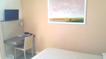 A hotel room with a bed, a desk with a chair, a television on the wall, and a painting.