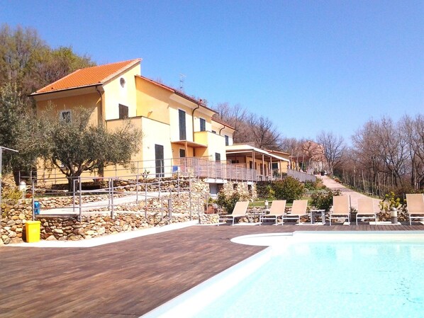A two-story building with a stone retaining wall and a pool area with lounge chairs. - Hotel Oroverde (Bergeggi)