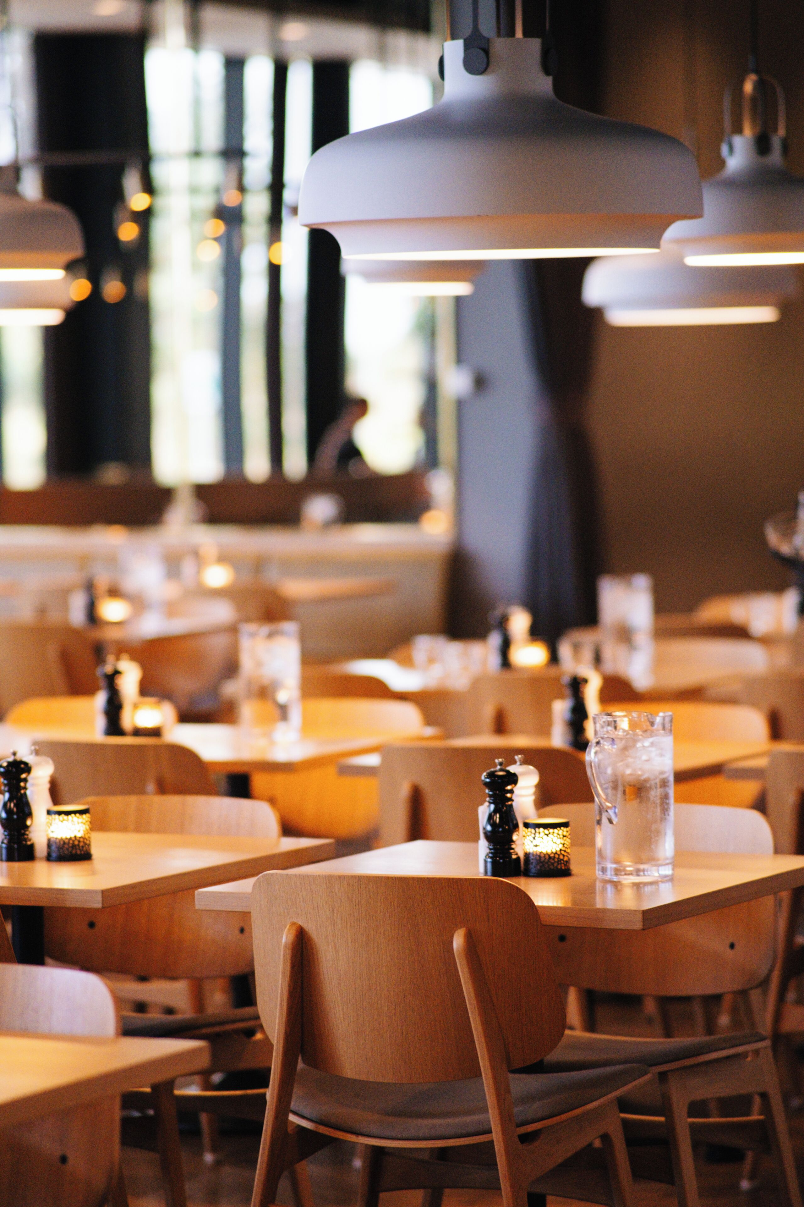 A restaurant interior with wooden tables and chairs, pendant lighting, and a glass pitcher on the tables.