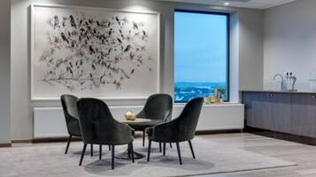 Un comedor moderno con un cuadro abstracto grande, vistas de la ciudad a través de una ventana y una cocina con armarios y un lavabo.
