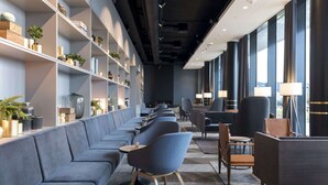 A modern lounge area with a long sofa, chairs, and tables, featuring a bookshelf with decorative items and plants. - Clarion Hotel Air (Sola)