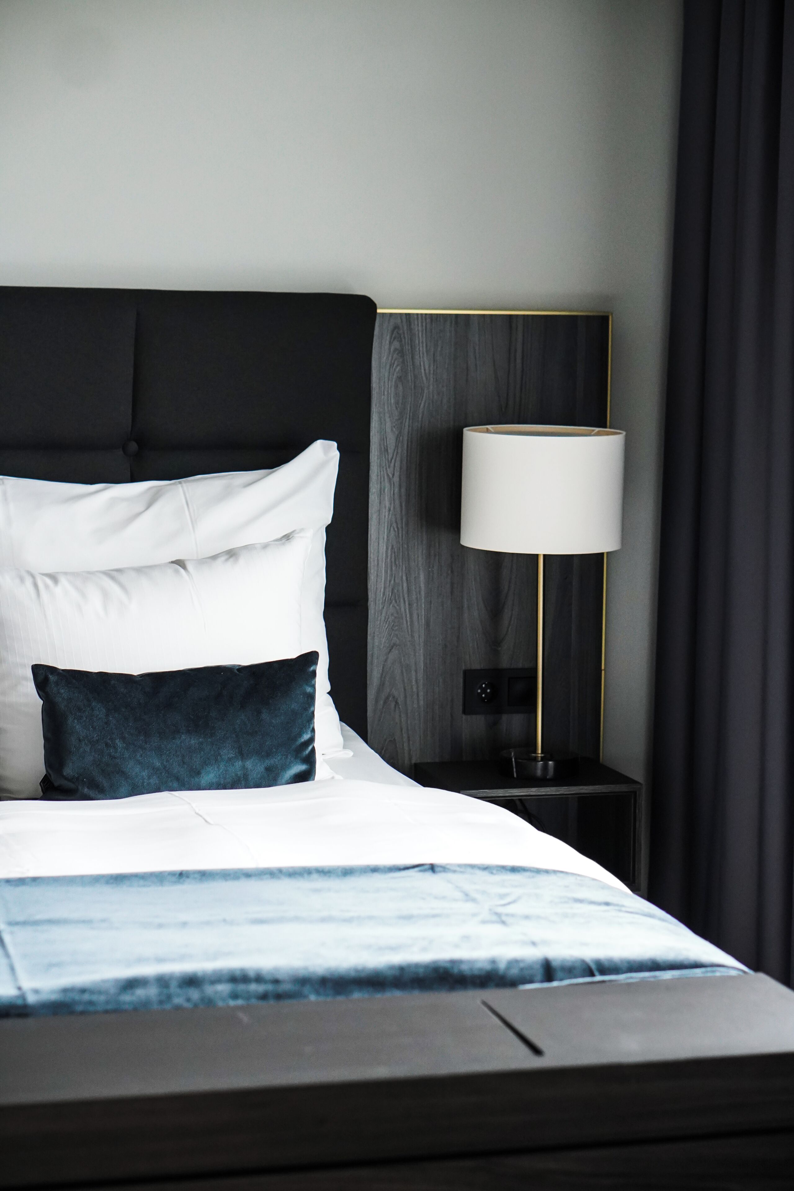 A neatly made bed with white and dark blue pillows, a bedside table with a lamp, and a dark headboard.