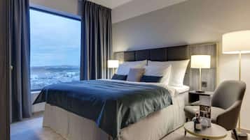 Habitación de hotel con una cama grande, vistas de la ciudad, una mesita con una botella y una silla.