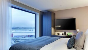 A hotel room with a large bed, a flat-screen TV, a bedside table with a lamp, and a view of a cityscape through a large window. - Clarion Hotel Air (Sola)