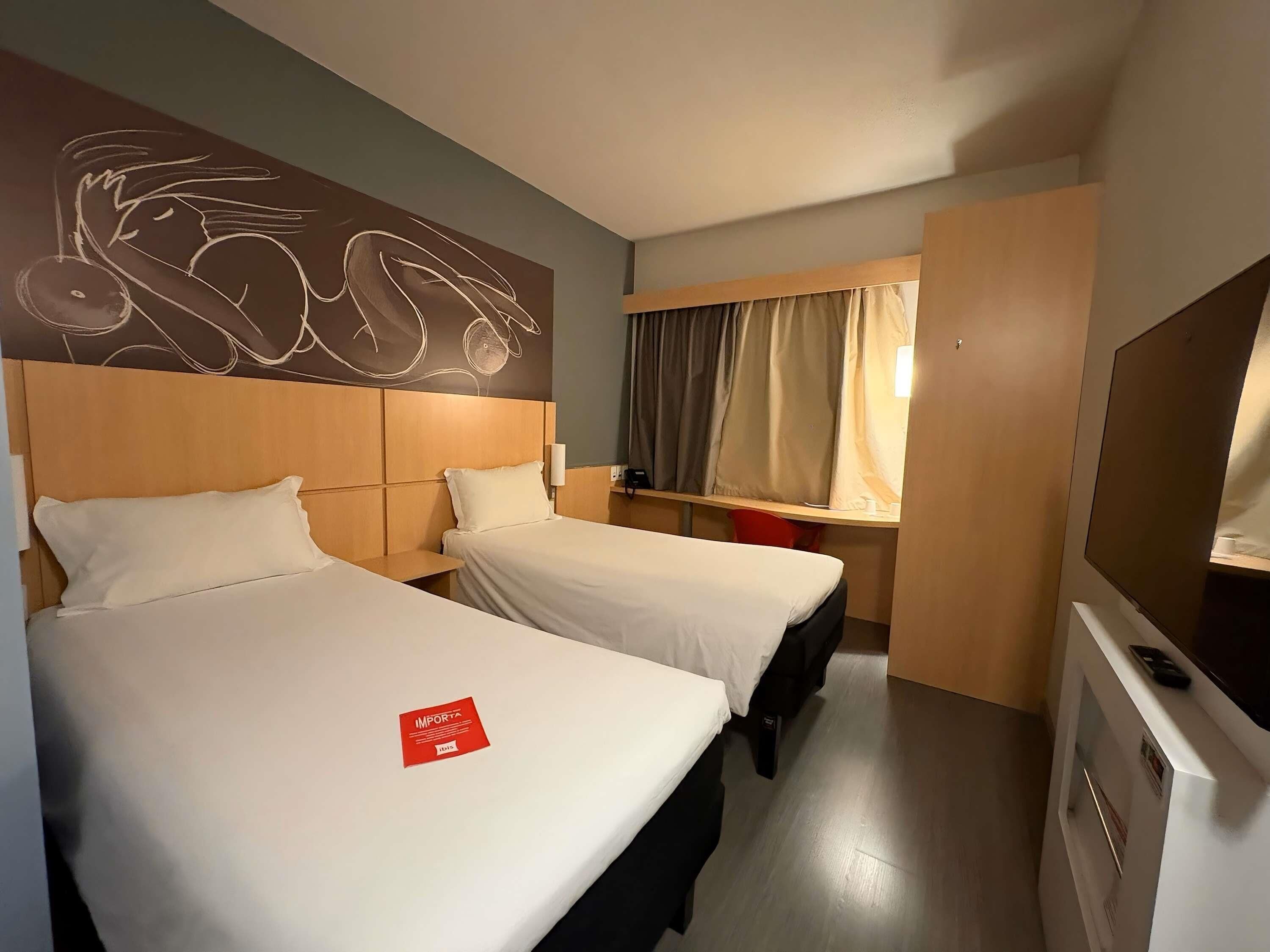 Habitación de hotel con dos camas, cabecero de madera, una obra de arte en la pared, escritorio con silla, ventana con cortinas y televisor.