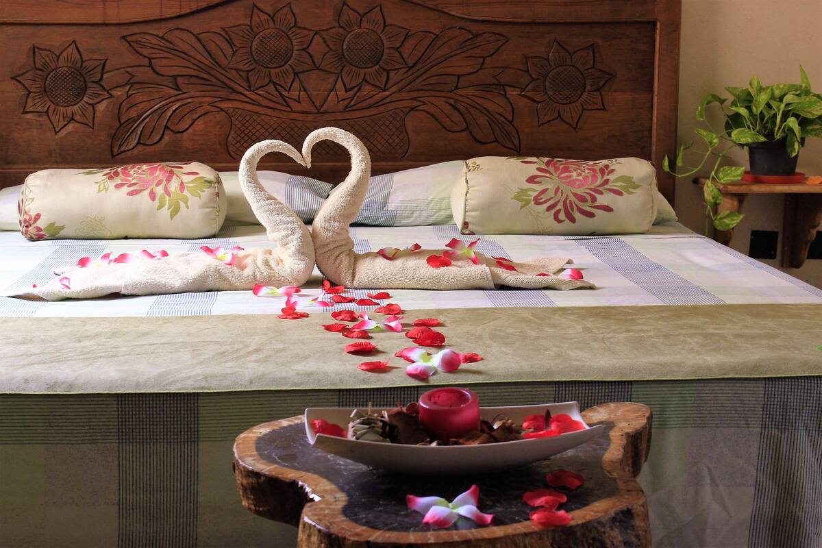A bed with floral pillows, a heart-shaped towel arrangement, and rose petals on the bedspread.
