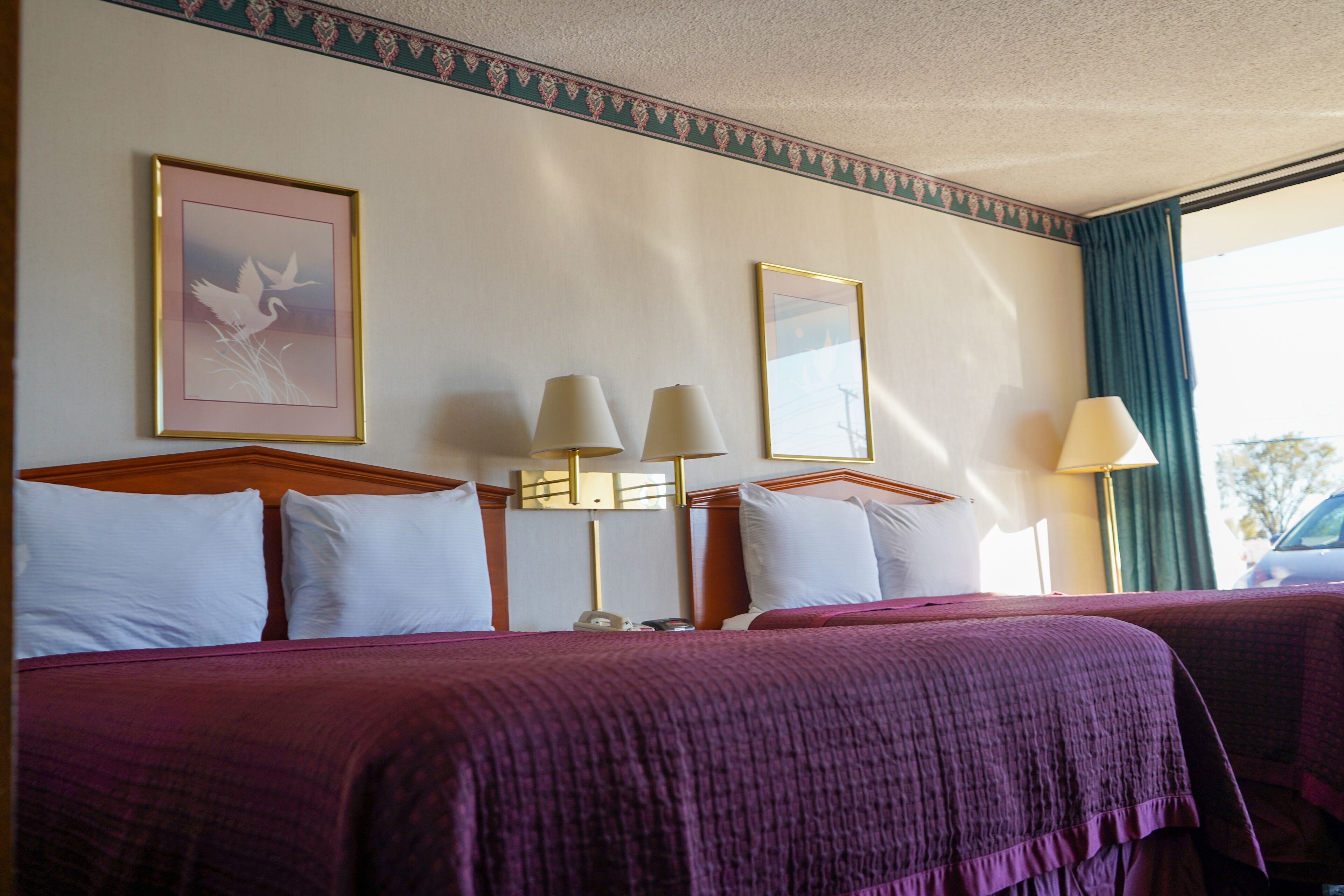 A hotel room with two beds, each with a headboard, white pillows, and a purple bedspread. There are two bedside lamps and a framed picture on the wall.