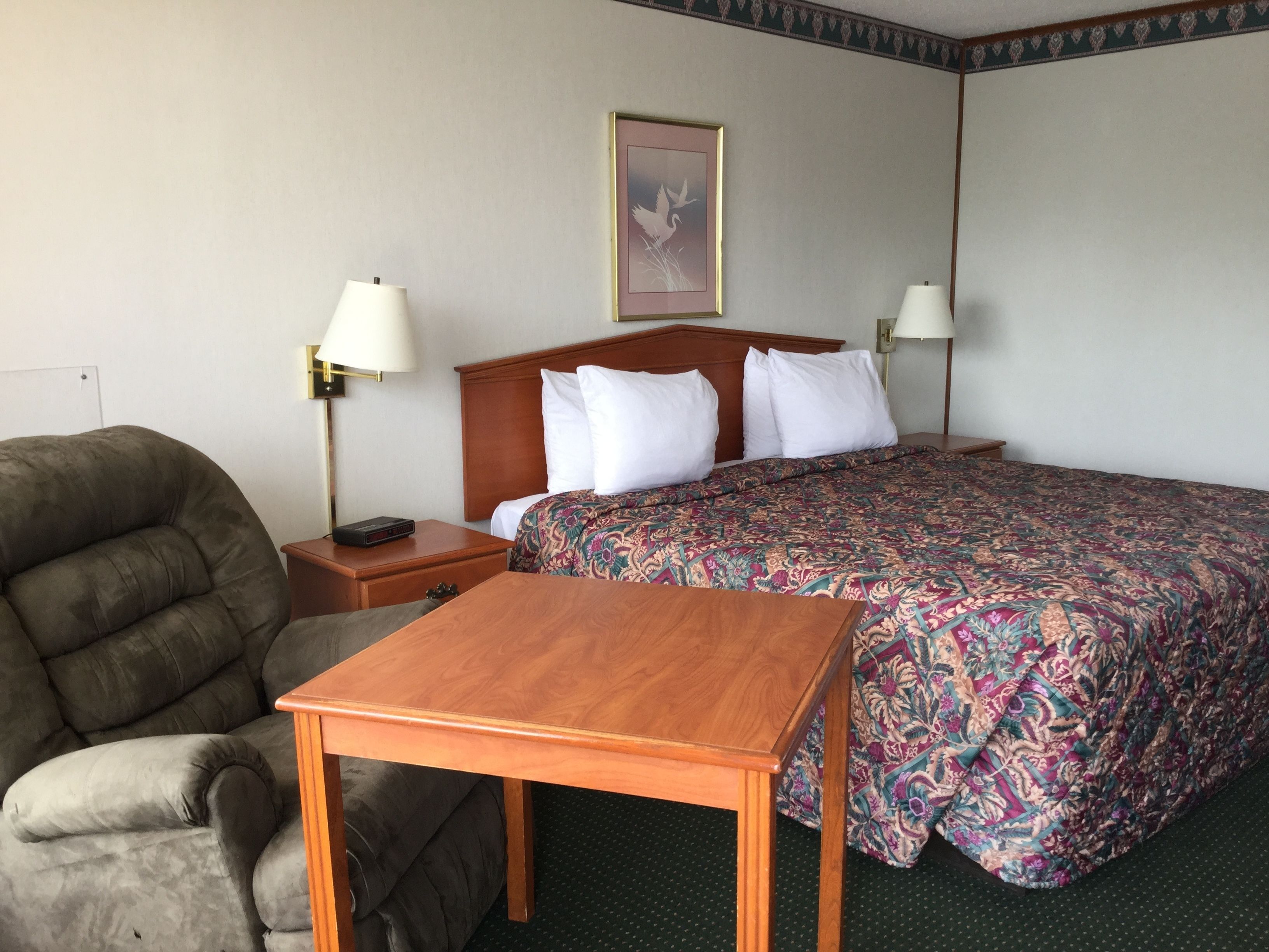 A hotel room with a large bed, two bedside tables, a wooden nightstand, a grey armchair, and a small table.