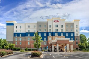 Ein mehrstöckiges Hotel mit einem gut sichtbaren Hampton Inn & Suites Schild, einem Parkplatz und einem begrünten Eingangsbereich.
