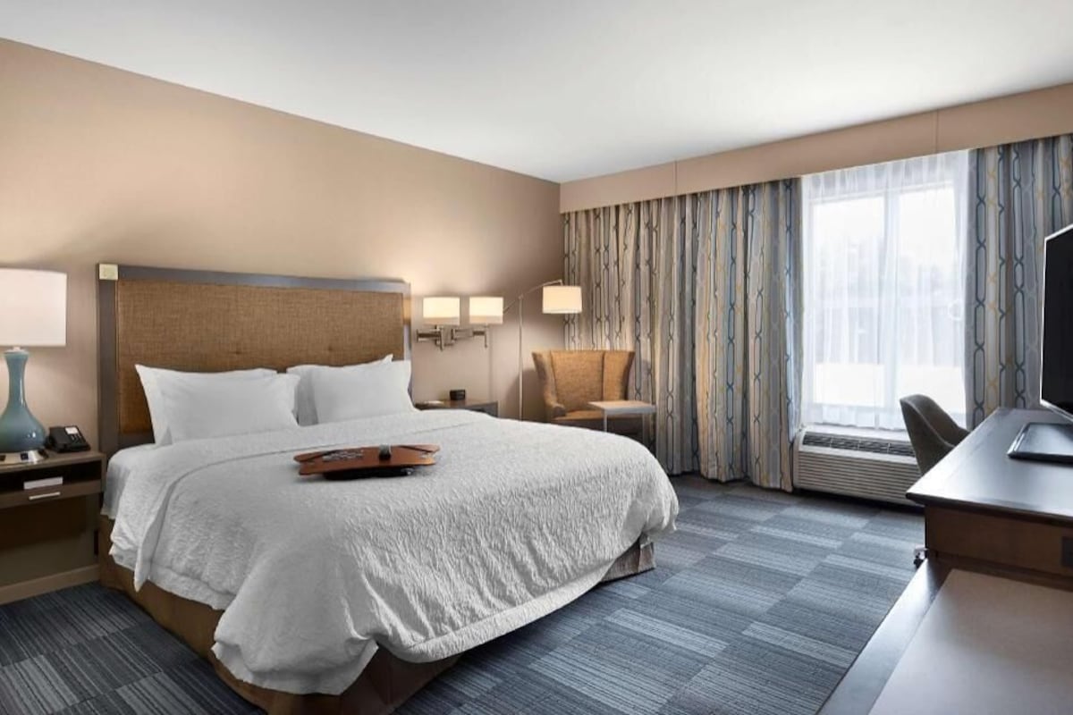 Ein Hotelzimmer mit einem großen Bett, einem Schreibtisch, einem Sessel, einem Fernseher und einem Fenster mit Vorhängen.