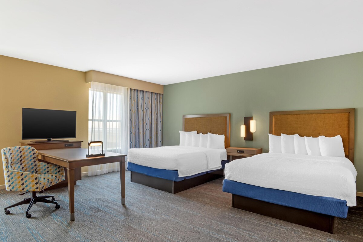 Ein Hotelzimmer mit zwei Betten, einem Schreibtisch, einem Sessel, einem Fernseher und einem Fenster mit Vorhängen.