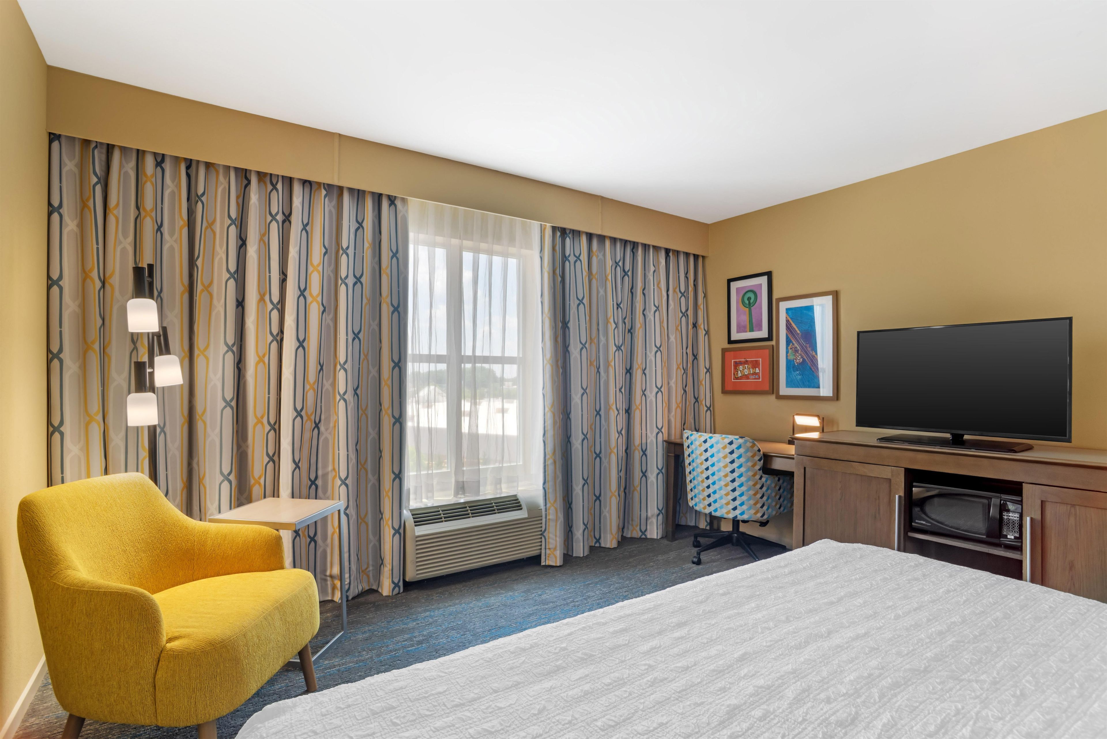 Una habitación de hotel con una cama, un sillón amarillo, un escritorio con televisor de pantalla plana, un microondas y una ventana con cortinas.