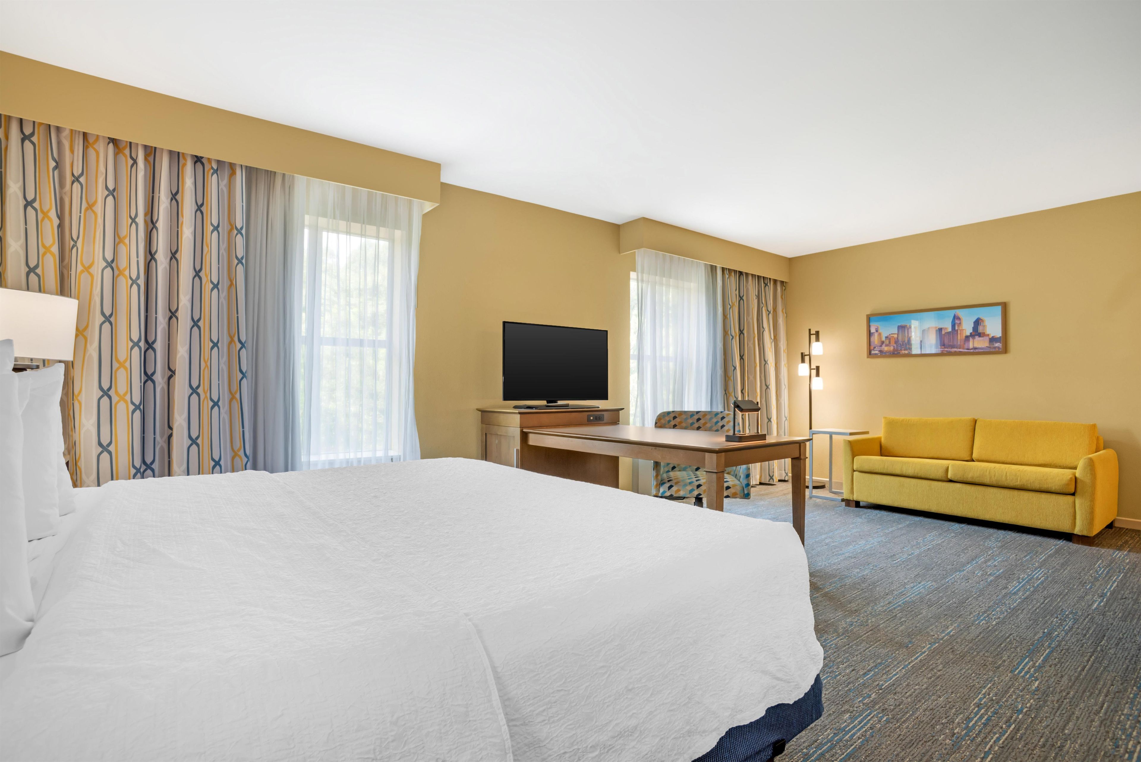 Habitación de hotel con una cama grande, un escritorio con televisor, un sofá y un cuadro en la pared.