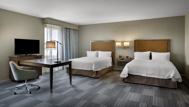 A hotel room with two beds, a desk with a TV, a chair, and a window with curtains.