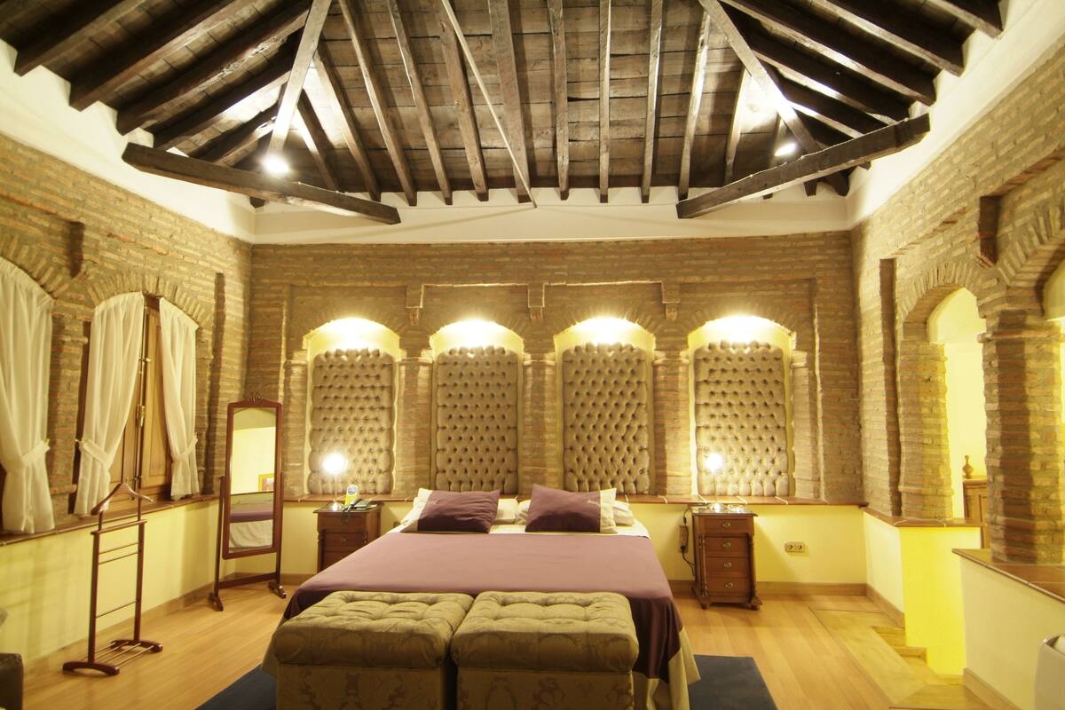 A bedroom with a wooden ceiling, brick walls, a large bed with a headboard, two bedside tables, and a mirror.