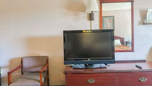 A hotel room with a television on a wooden dresser, a chair, and a mirror reflecting a bed and a wall-mounted lamp.