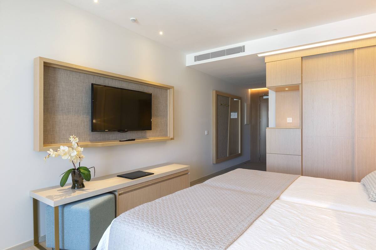 A modern hotel room with a large bed, a flat-screen TV mounted on the wall, a small desk with a chair, and a walk-in closet.