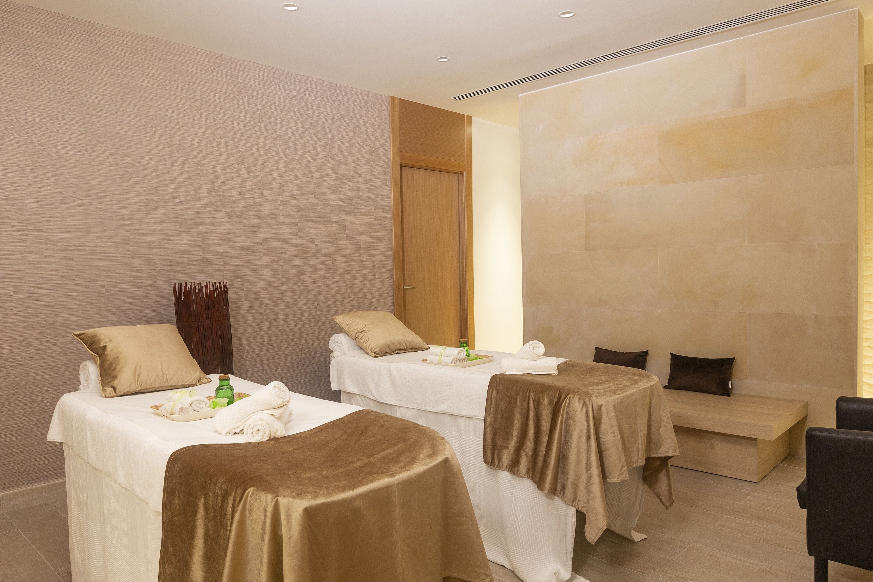 Habitación de hotel con dos camas, cada una cubierta con un edredón marrón, un banco de madera y una mesita de noche con artículos de tocador.