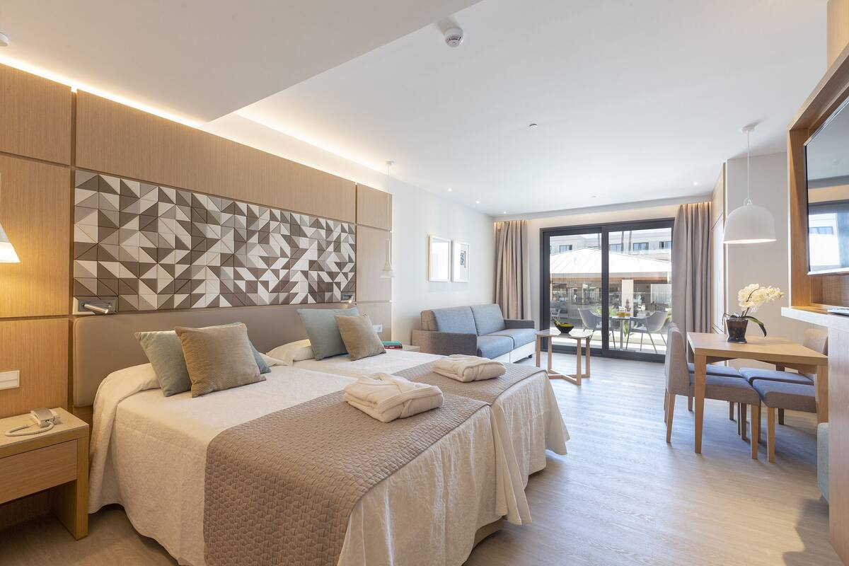 A modern hotel room with a large bed, a sofa, a dining table, and a view of the city.