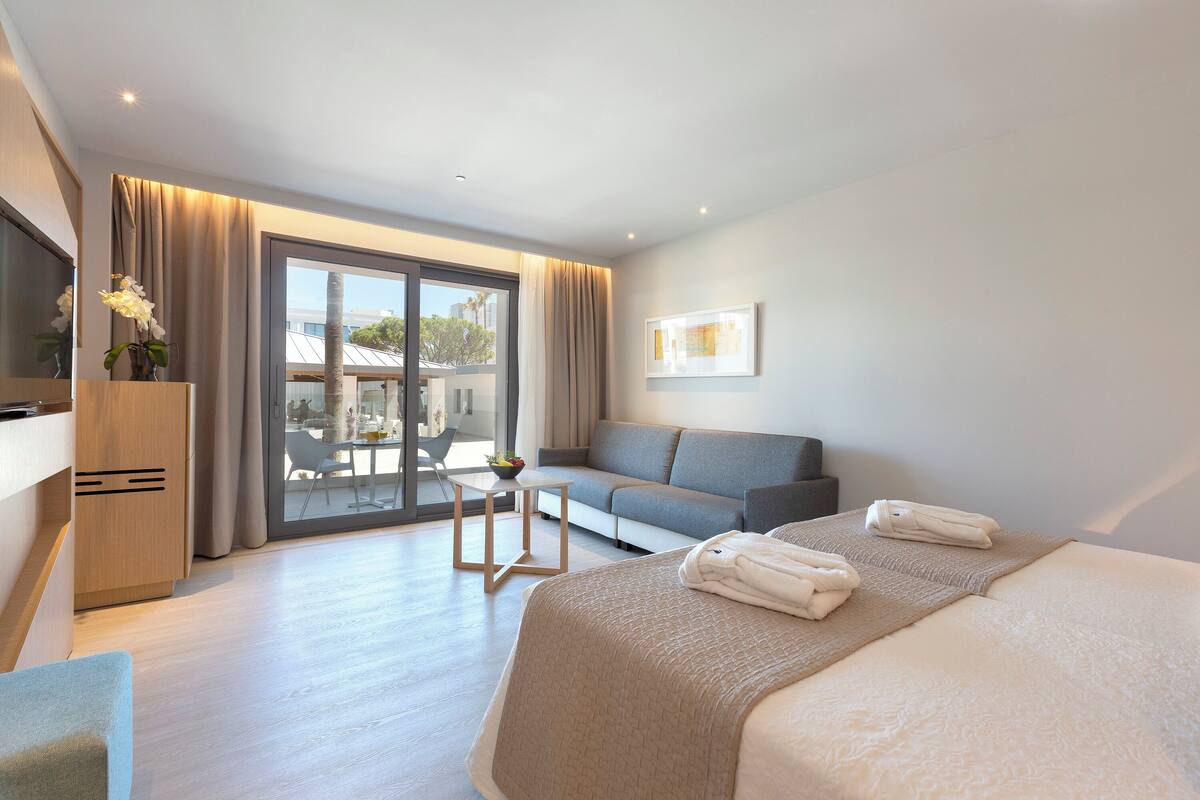 A modern hotel room with a large bed, a sofa, a small table, and a view of the outdoors.
