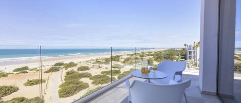 A balcony with a glass railing overlooking a beach and ocean. There is a table with chairs and a glass of orange juice.