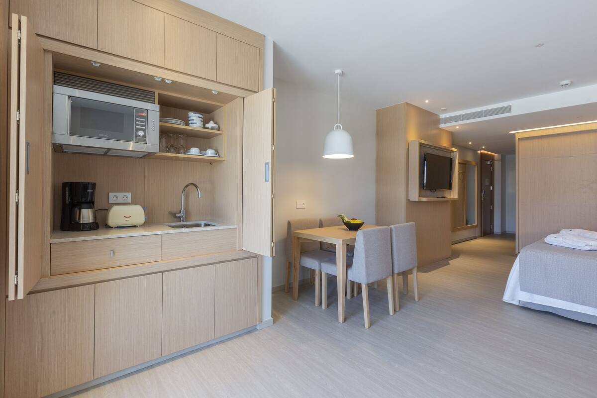 A modern hotel room with a kitchenette, a dining area, a bed, and a TV.