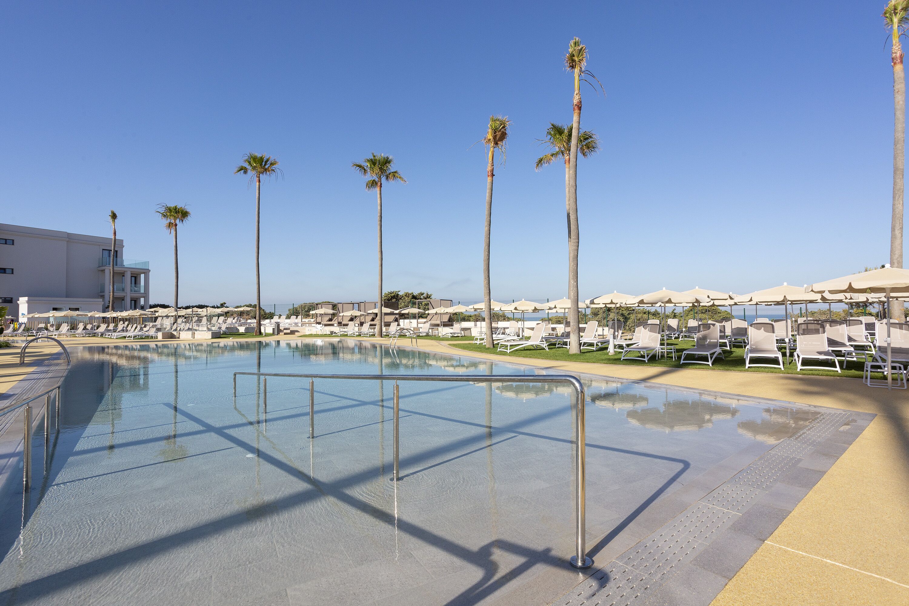 Piscina con sillas de descanso y palmeras, rodeada por un edificio de diseño moderno.