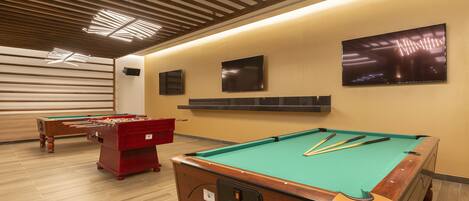 A pool table with cues, a foosball table, and a billiards table in a room with wooden flooring and a slatted ceiling.